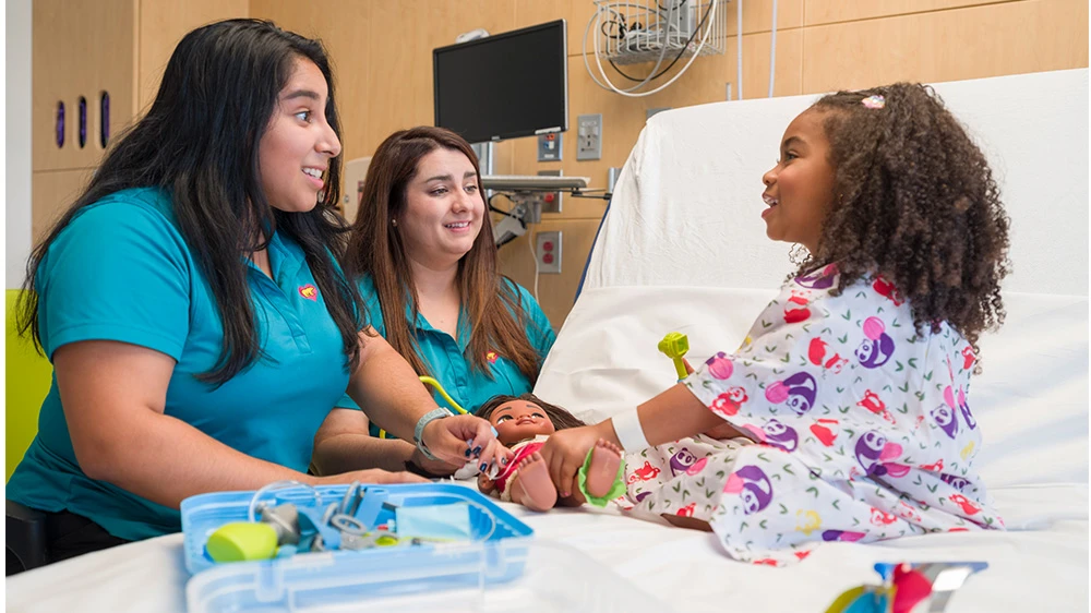 2 child life specialists playing games with girl at hospital bed.